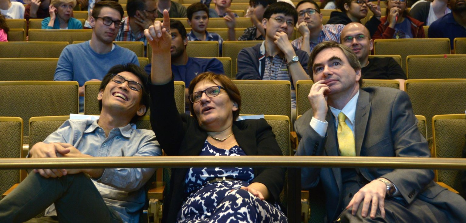 Members of the audience try to figure out the puzzle given by Burger. One woman points at the screen, while another laughs.