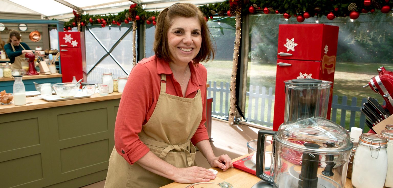 Tina Zaccardi, wearing a beige apron and a dark pink top, smiles for the camera in a kitchen.