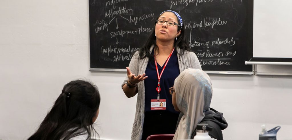 A standing woman speaks and holds up her right hand in front of a chalkboard