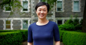 A smiling Asian woman wearing a blue dress and standing in front of a building