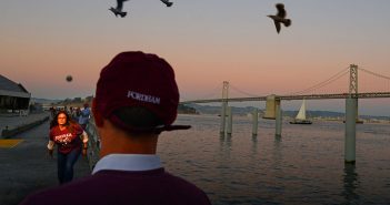 Fordham Law graduate Christina Chiaramonte practices bocce throws outside the Ferry Terminal Building on San Francisco’s Embarcadero at dusk in October 2019, with three birds flying in the sky above and the Golden Gate Bridge and a sailboat visible in the background.