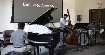 Three men each play an instrument: a piano, a cello, and a drum set, in a room with white walls.