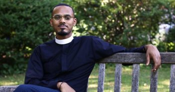 A man wearing glasses and black priest robes sits on an outdoor bench and smiles at the camera.