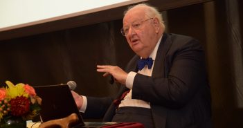 Nobel Laureate and Renowned Economist Addresses Global Poverty Issues at Fordham Conference A seated elderly man wearing a suit and a blue bow speaks in front of a black curtain.