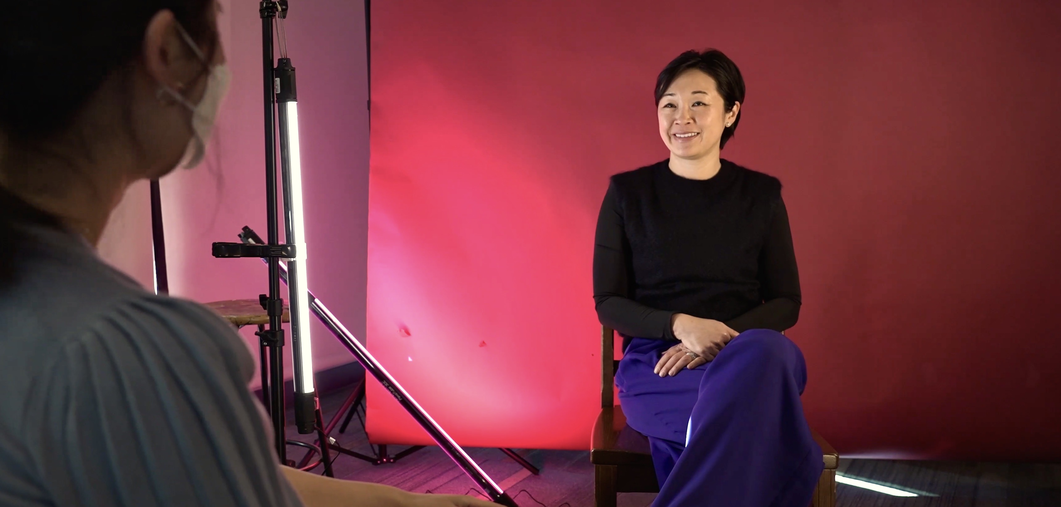 An Asian woman smiles against a fuchsia backdrop while speaking to another person.