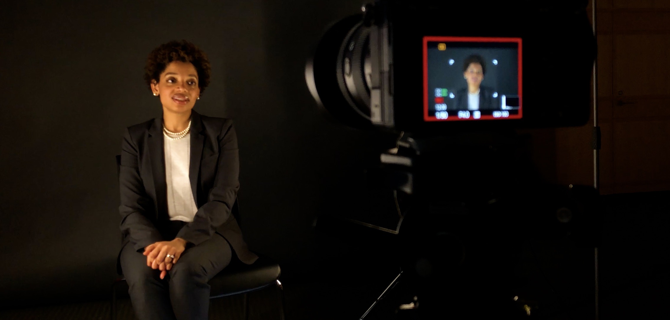 Teaching Theology to Women in Prison A woman sits in front of a black backdrop with a camera pointed towards her.