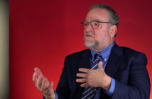 A man gestures with his hands while sitting in front of a red backdrop.