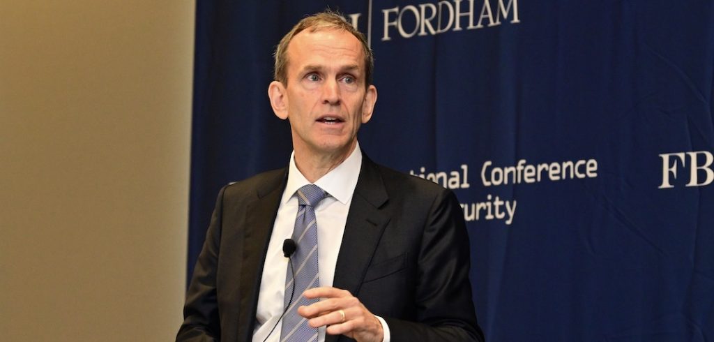 A man wearing a suit, a tie, and a lavalier mic clipped to his tie speaks in front of a blue wall.