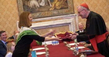 A woman gives a maroon baseball cap to a man wearing a priestly outfit.