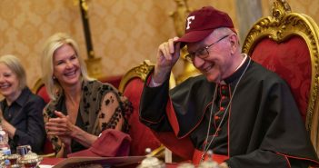 A man wearing a maroon baseball cap and a black priestly outfit smiles.