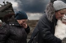 Two women, one holding a white cat in her arms, the other covering her mouth with her hand, flee a battle scene in Irpin, Ukraine