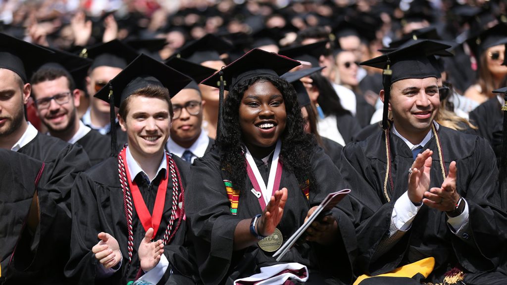 Students in caps and gowns sitting and clapping. Smiling.