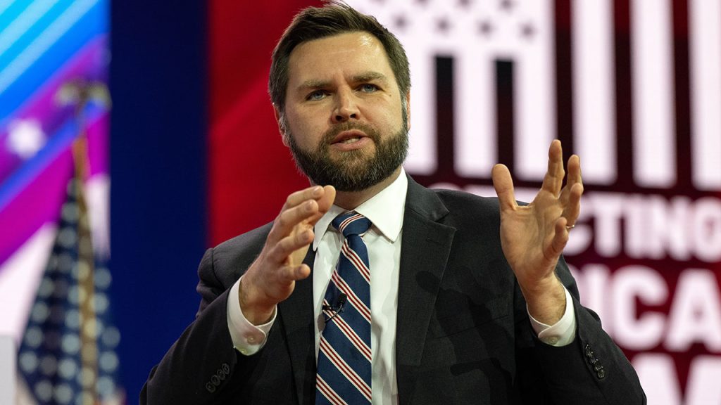 A man with a beard wearing a tied speaks whole holding hands up in the air.