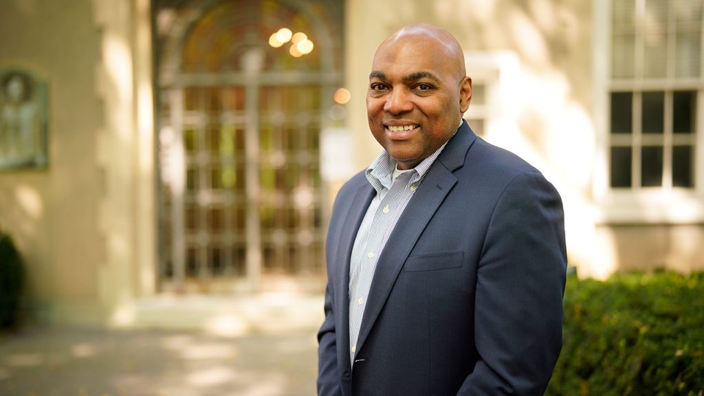 New Vice President for Equity and Inclusion Sees Strength in Differences A man with a suit standing in front of a building with a door in the background