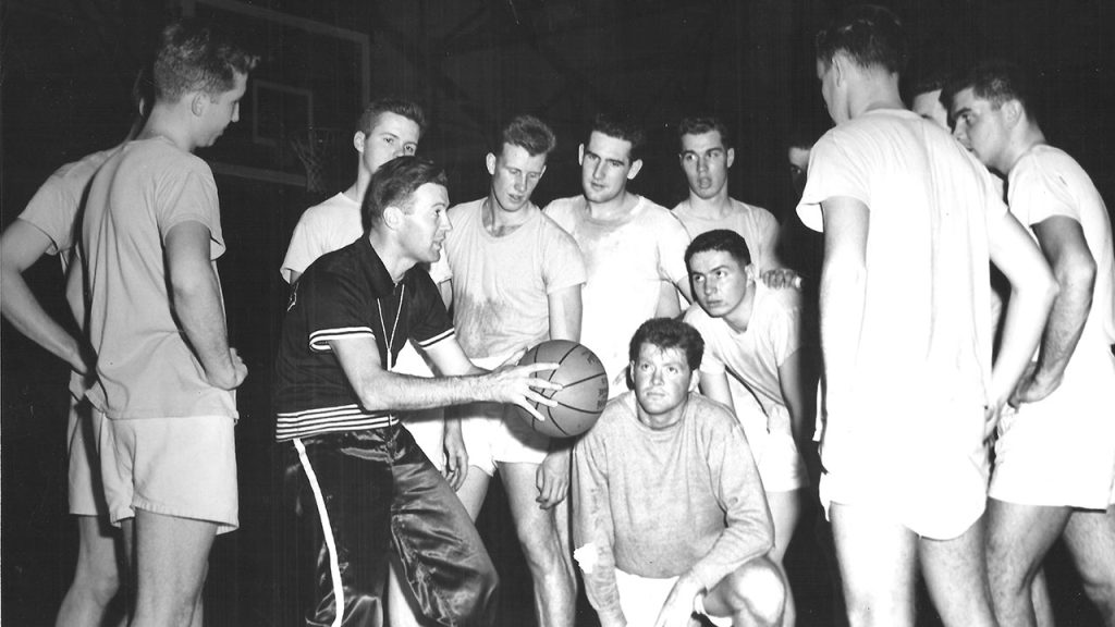 Editor’s Note: Coach Johnny Bach and the Art of Elevation Basketball players gather around and look at a coach holding a basketball in the middle of the huddle