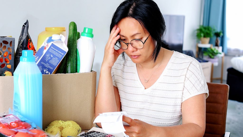 Stress over Inflation Increased Even After Prices Cooled, Study Shows A woman seated at a table with groceries, looking anxiously at her receipt