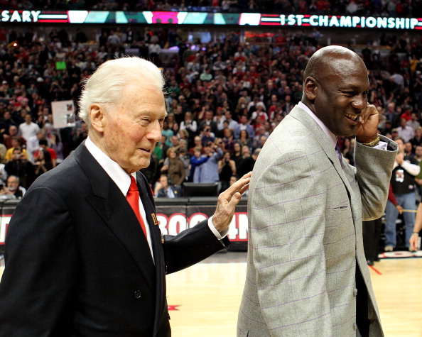 Former Bulls assistant coach Johnny Bach puts his hand on his former player Michael Jordan's back as the pair, dressed in suits, walk across a basketball court to the cheers of fans