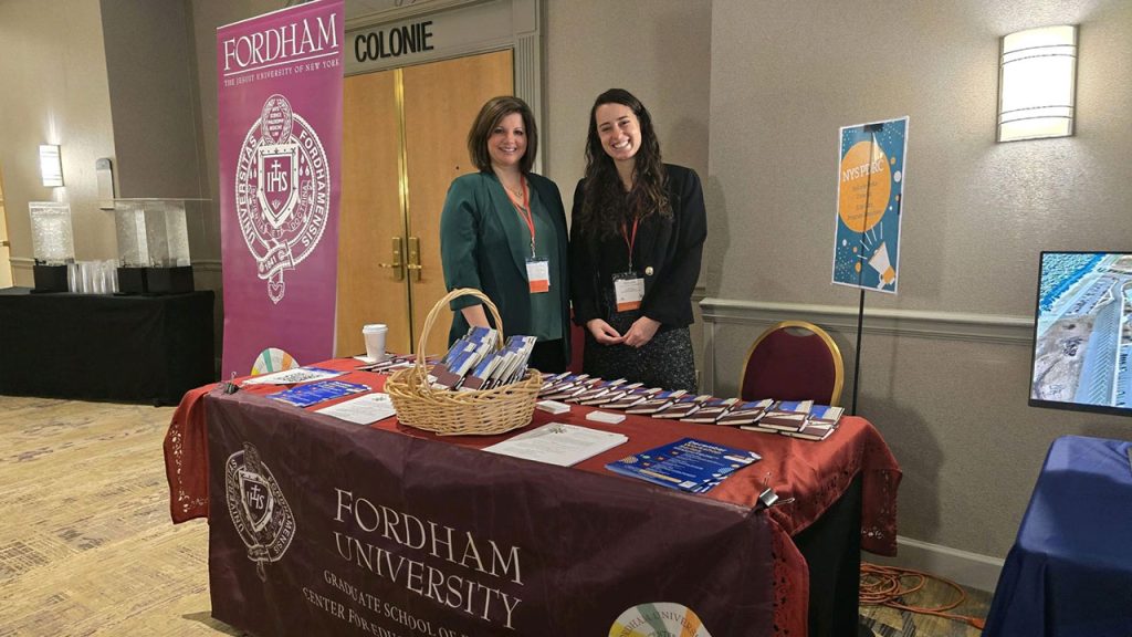 Two women standing behind a table with a Fordham banner behind them.