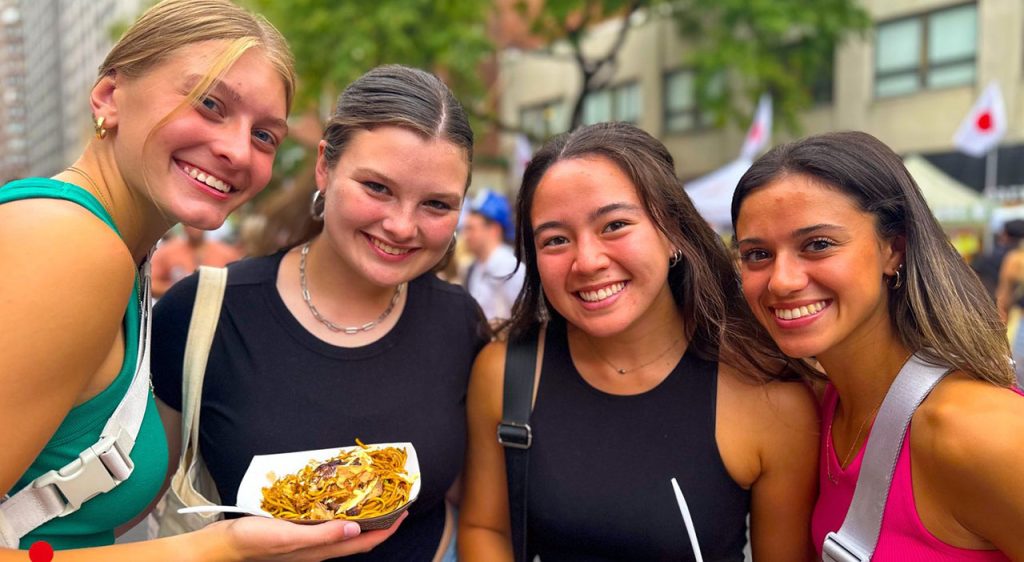 4 women smiling together, one on left holding noodles