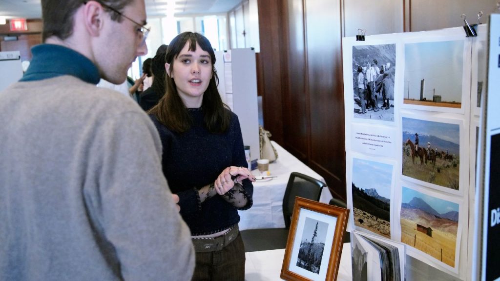 Fordham student Alison Kulak stands in front of a poster board chatting with a male about her project during the ARS Nova Arts and Research Showcase.