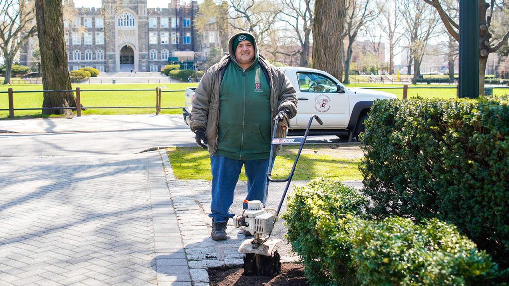 A man wearing a heavy hoody and jeans stands behind a tiller perched in a mound of black dirt.