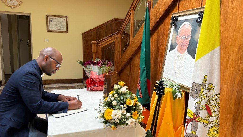 A man in a priest's uniform sits at a table signing a book before a photo of Pope Francis