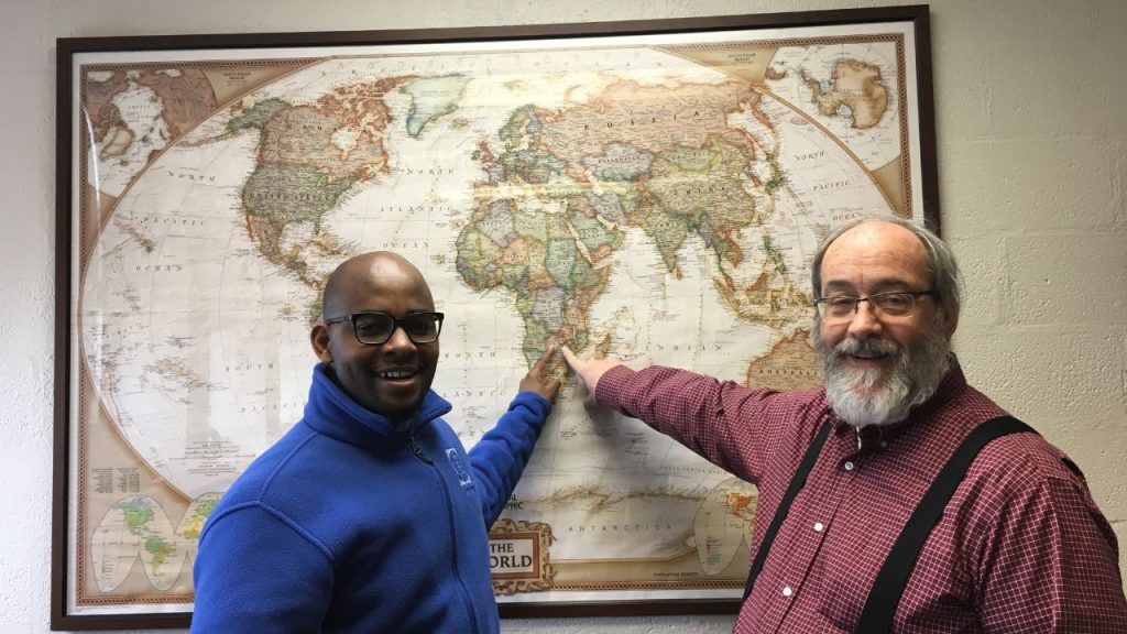 Two men stand side by side pointing to Zambia on a map of the world