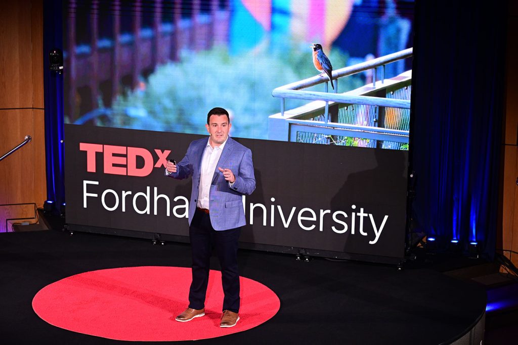 Dustin Partridge speaks from stage in front of an image of a robin perched on a balcony is shown behind him.