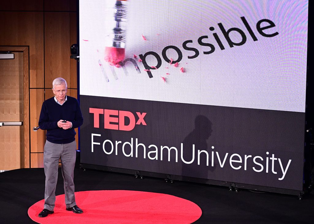 David Marcotte speaks from stage while the image of the word "impossible" is shown having the  "im" part erased by a pencil eraser.