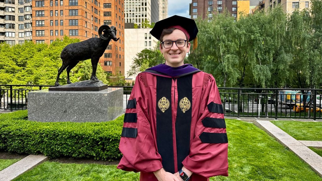 Timothy Bouffard, in cap and gown posing near Fordham ram statue on Lincoln Center Campus