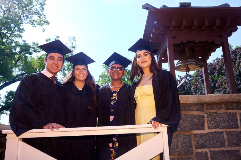 Two men and two women in caps and gowns next to the victory bell 