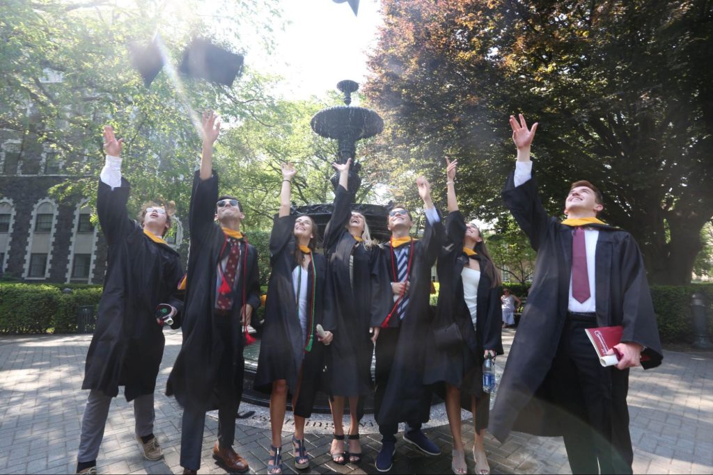 Group of men and women in gowns throwing caps in the air in front of a fountain 