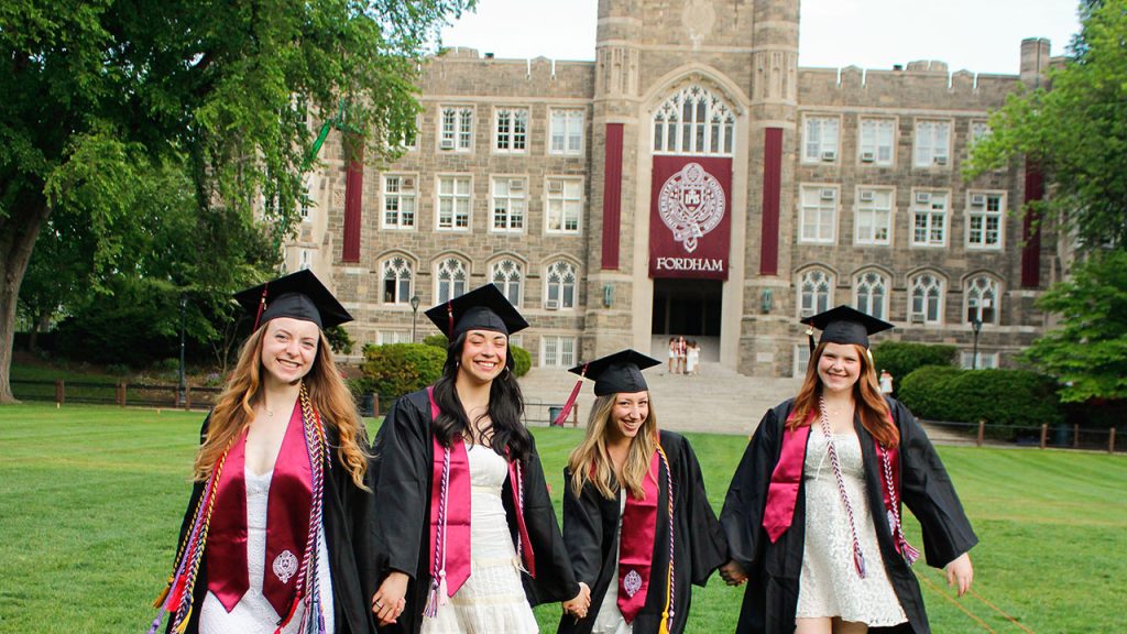 4 women in caps and gowns, smiling holding hands on Edwards parade. Keating Hall in background.