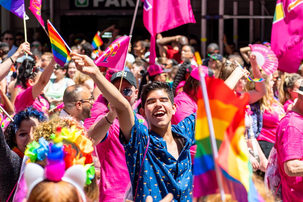 Smiling male presenting individual in crowd of people at pride parade