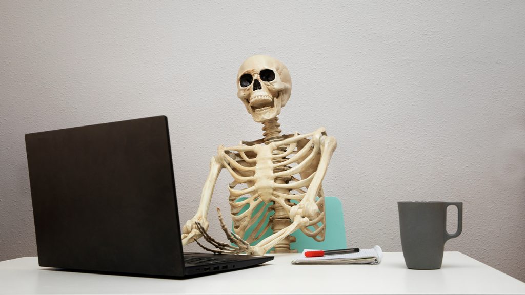 A skeleton sits at a desk with a laptop and mug of coffee