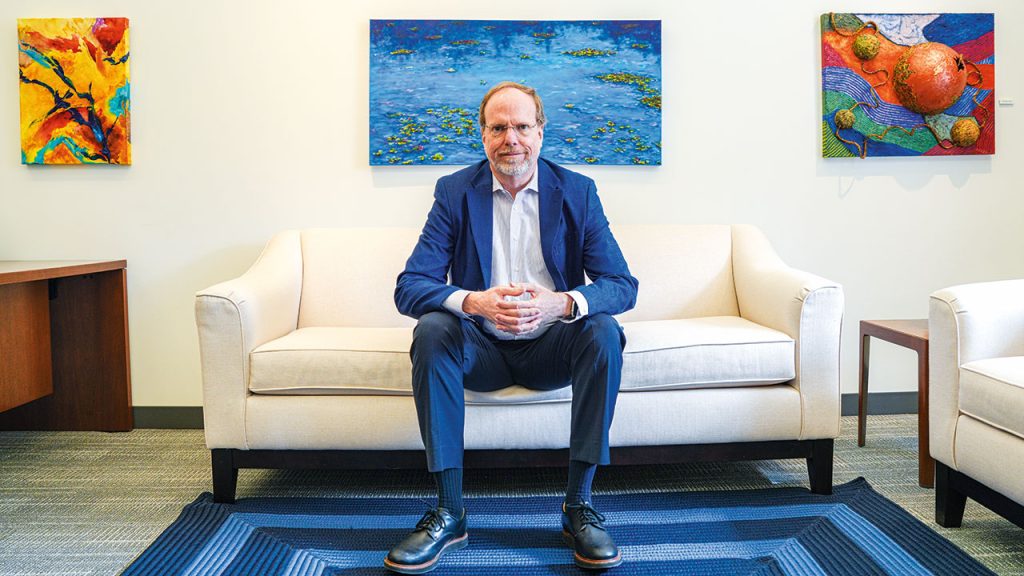 Dennis Jacobs wearing blue suit and white shirt sits on off-white couch in front of three paintings