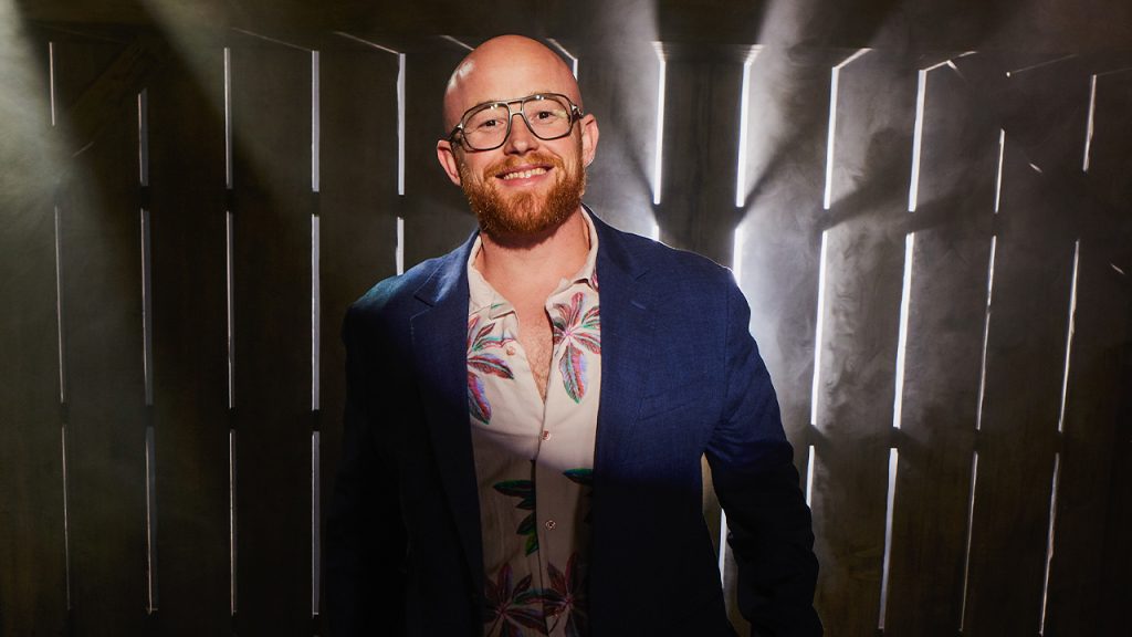 portrait of Craig Calefate wearing jacket and tropical shirt in front of wooden fence with dramatic smoke.