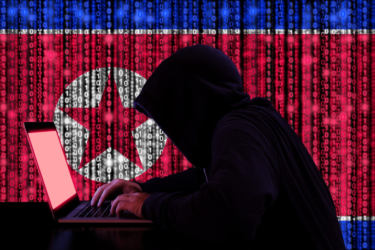 A man in a hoodie works on a labtop in front of a digitized image of the North Korean flag, representing the North Korean remote IT worker scam discussed at Fordham's ICCS