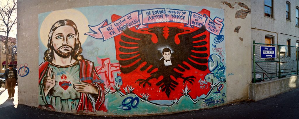 A panoramic memorial mural painted on a light-colored building wall. On the left is a portrait of Jesus Christ with a halo and a Sacred Heart on his chest. The center features a large black double-headed eagle, the national symbol of Albania, painted on a red field. In the center of the eagle is a portrait of a man with dark hair and a mustache, dressed in a tuxedo. A banner above the eagle reads, 'IN LOVING MEMORY OF ANTON D. MIRCA' and 'NE RROFTË TE PA HARRESUAR.' Smaller text on the right says, 'REST IN HEAVEN' and 'WE'LL MISS YOU!.' The mural is painted above a strip of simulated green grass and is located on a street corner next to a chain-link fence