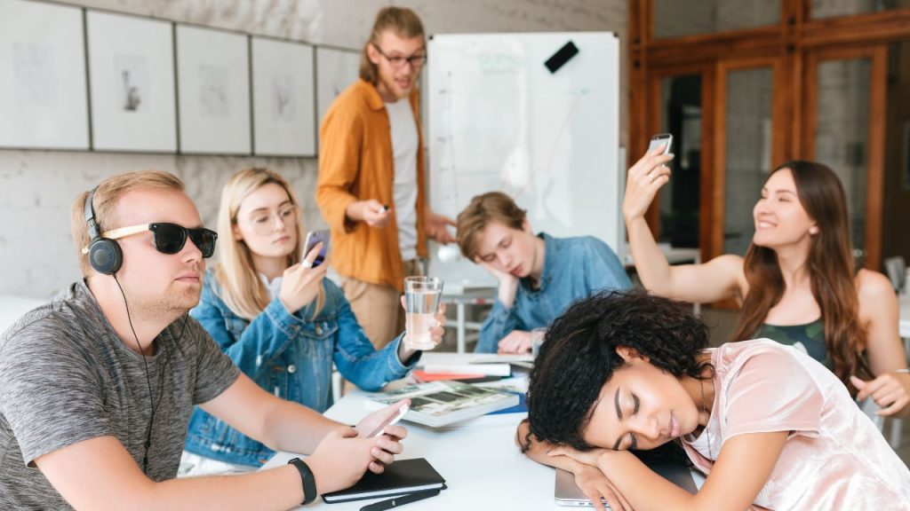 A group of students sitting around a white board looking unfocused while a group leader looks frustrated representing group project challenges