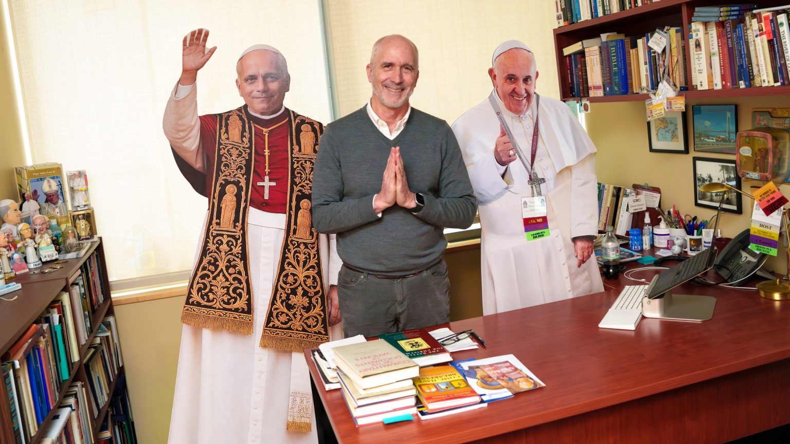 David Gibson stands, smiles, and holds his hands in prayer in between life-sized cardboard cutouts of Pope Leo and Pope Francis.