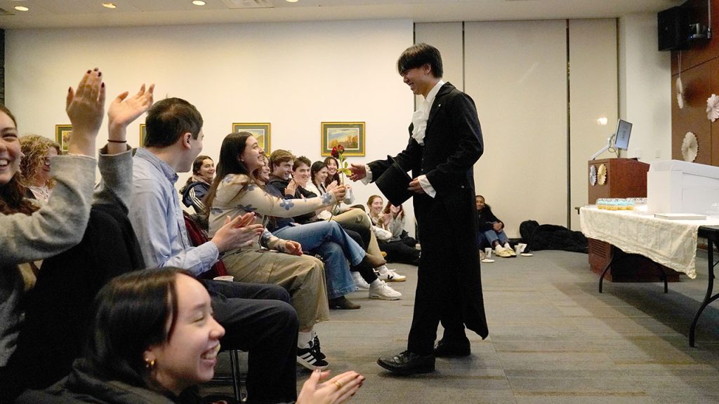 A man wearing a tuxedo hands a rose to students seated in the audience